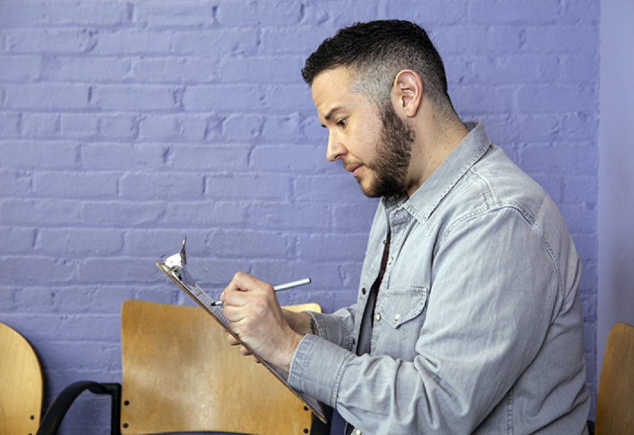 A transgender man sits in a waiting room, filling out paperwork on a clipboard.