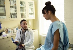A transgender woman in a hospital gown speaking to her doctor.